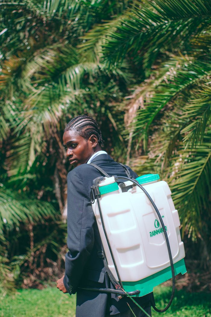 A man in a suit carrying a pesticide sprayer outdoors among palm trees.